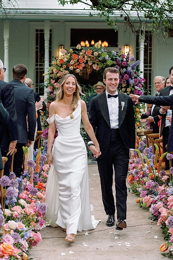 Wedding recessional as bride and groom walk hand in hand down a flower-lined aisle, guests cheering in an outdoor courtyard setting