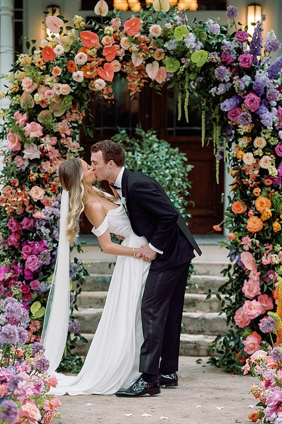 Wedding kiss portrait of bride and groom dipping under a colorful floral wedding arch with roses and greenery on stone steps by a doorway