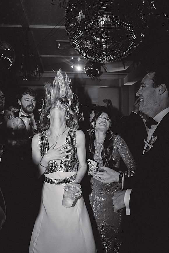 Wedding dance floor packed with wedding reception dancing guests in tuxedos and a sequin dress under a disco ball and chandelier lighting