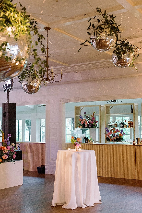 Wedding reception bar setup with hanging disco balls and greenery garlands, wood front shelves of bottles, and a candlelit cocktail table nearby