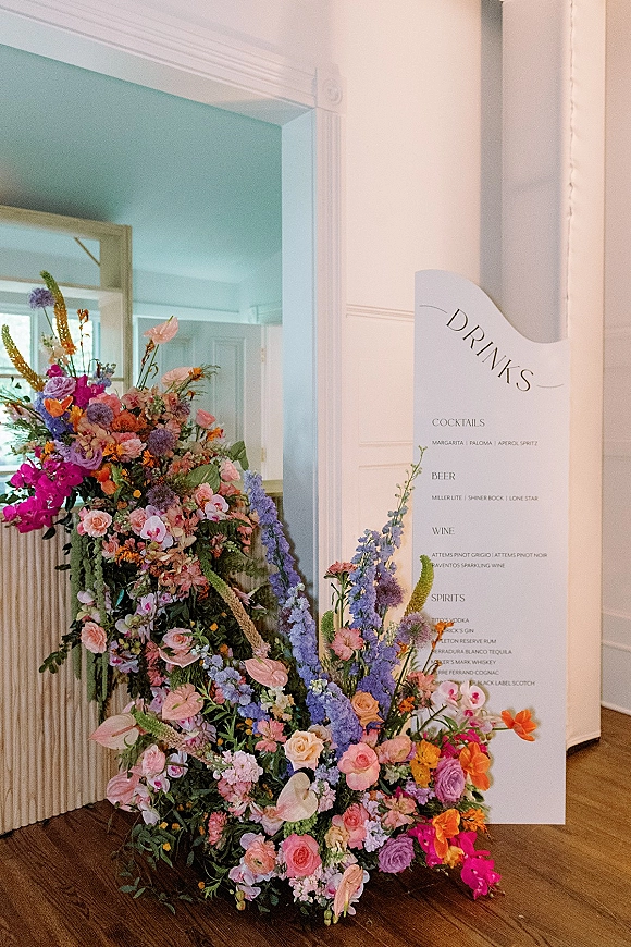Wedding bar signage with a wedding drink menu sign beside colorful wildflower accents, set against a white paneled wall indoors