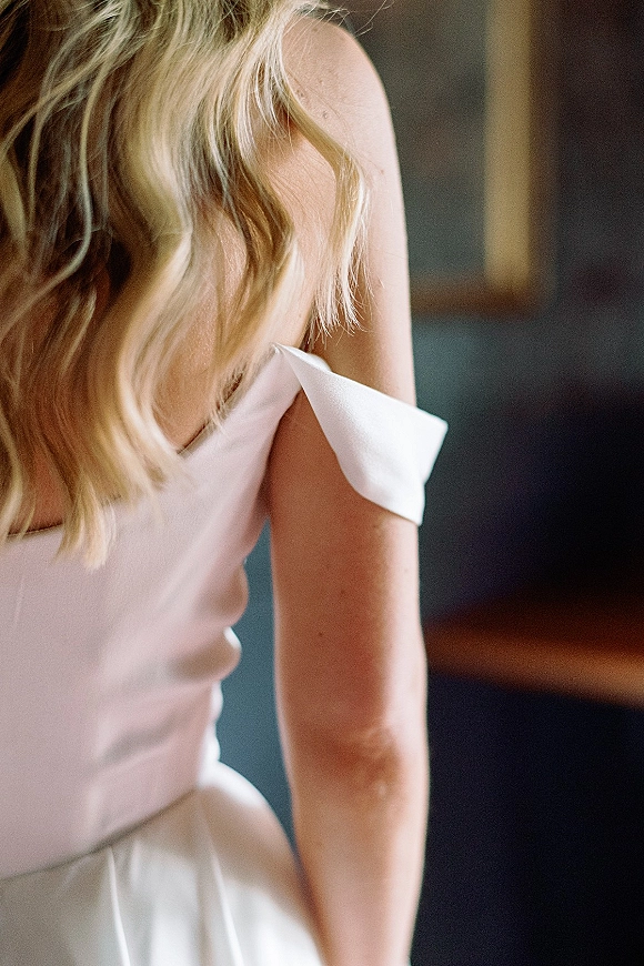Wedding dress detail with off the shoulder wedding dress neckline in smooth satin, softly lit by window light in a blurred indoor room
