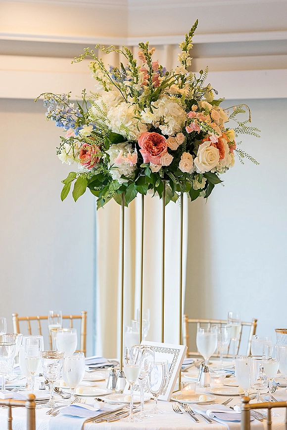 Reception centerpiece with tall wedding centerpiece of roses and hydrangea on a gold stand, above a formal place setting with votive candle in a paneled reception room