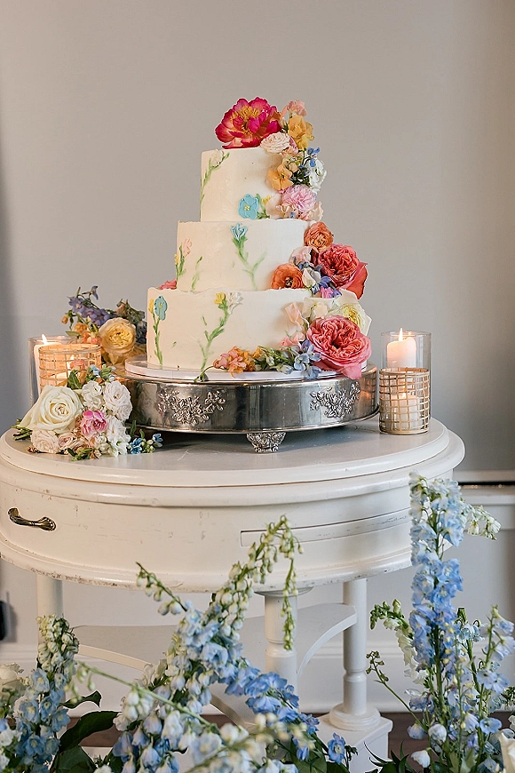 Wedding cake with three-tier buttercream finish, fresh flower accents on a silver stand, flanked by glass candle holders indoors
