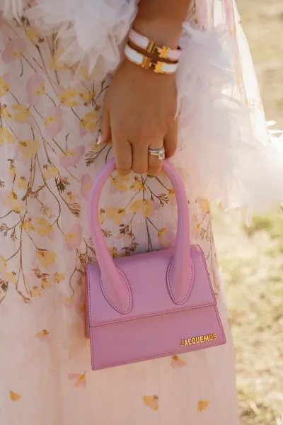 Wedding guest accessories with a lavender mini handbag, emerald-cut engagement ring, and gold-and-white bracelet stack in sunlit lawn setting