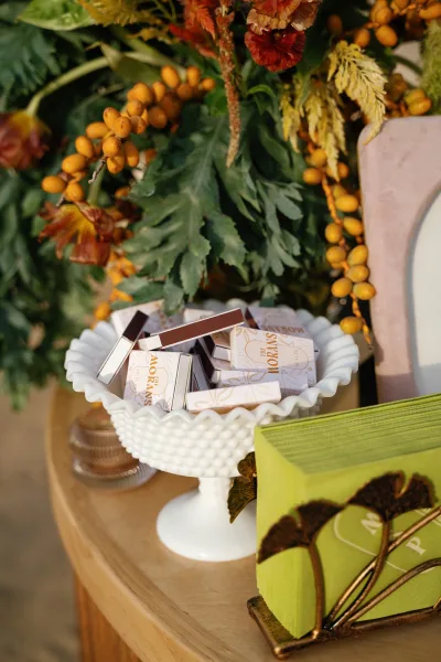 Wedding favors in petite boxed candy packages arranged in a pedestal bowl beside marigold and rust florals on a wood tabletop