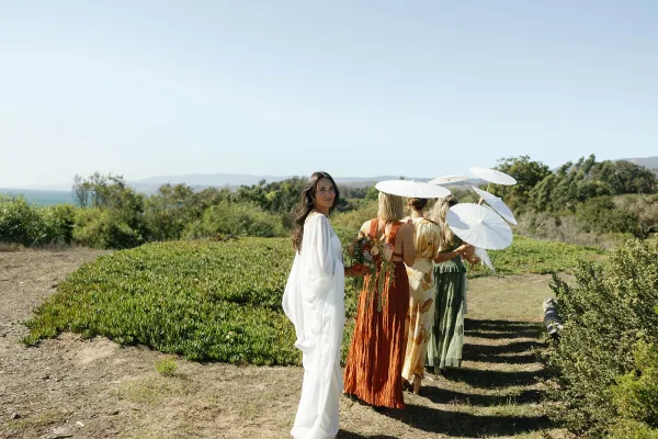 Bridal party portrait with bridesmaids with parasols, bride in white gown holding bouquet in a grassy field with mountains beyond