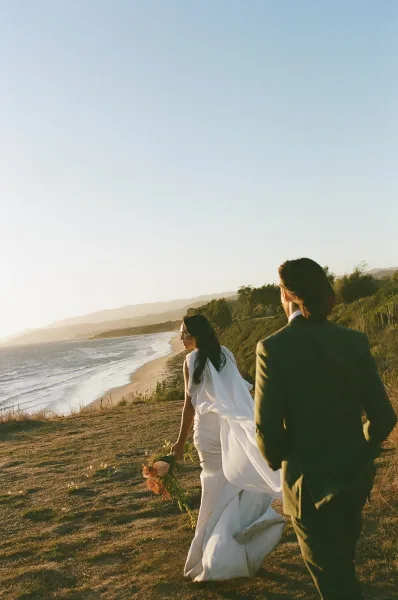 Couple portrait of bride and groom walking away, veil blowing as she holds a bouquet along a beach cliff path with ocean view