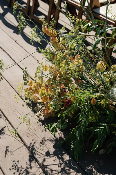 Aisle florals with ceremony aisle flowers in wildflower style greenery and seed pods on a wooden deck, sunlit beside ceremony chairs