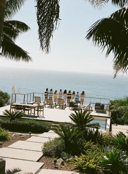 Bridal party photo of bridesmaids in striped robes on a patio deck, facing the ocean view with palm trees and coastal hillside beyond
