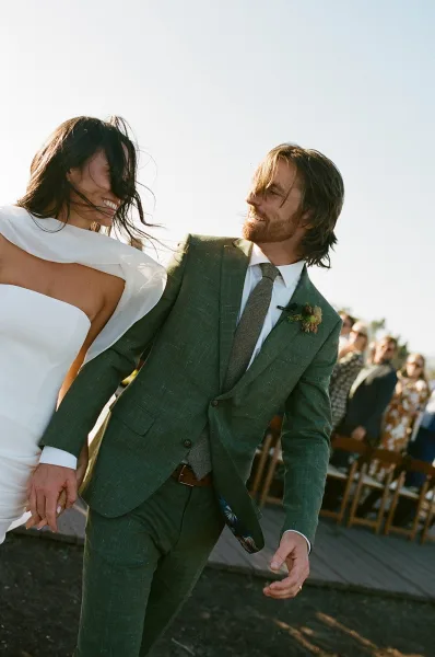 Wedding recessional as bride and groom walking hand in hand, bride laughing in a strapless dress while guests cheer along the outdoor aisle