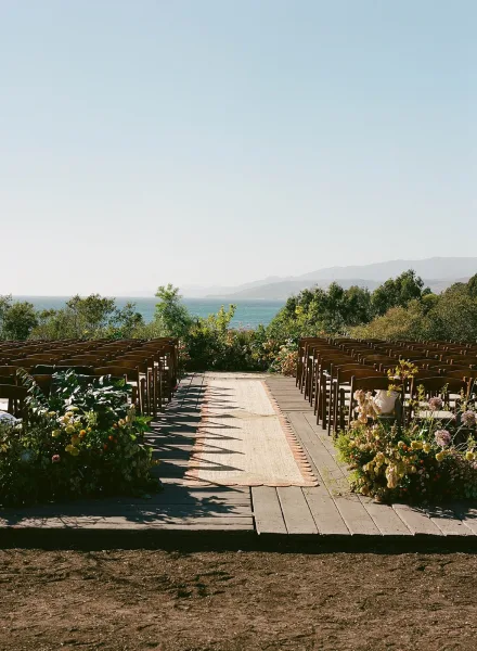 Ceremony setup with outdoor ceremony seating in wood chairs, neutral aisle runner and floral greenery on a deck overlooking the ocean coastline
