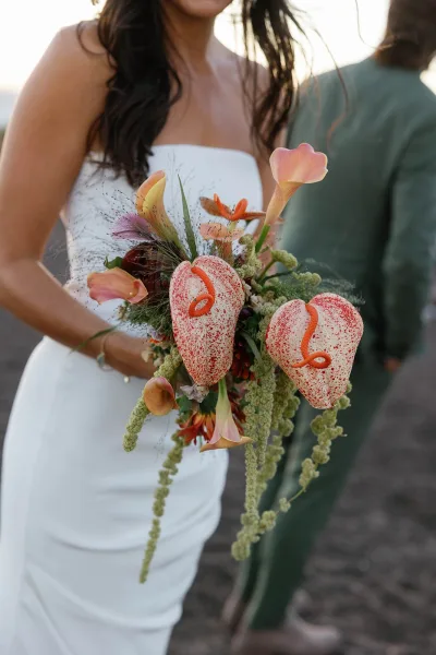 Bridal bouquet featuring anthurium wedding bouquet with calla lilies and trailing amaranthus, held by a bride in a strapless dress outdoors