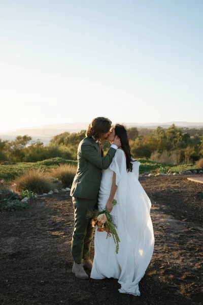 Wedding kiss portrait of bride and groom kissing as her long veil blows at sunset, with hills and trees behind on a dirt path