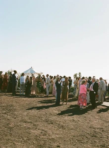Wedding cocktail hour guests mingling outdoors with drinks and sunglasses, suits and floral dresses beside a coastal ocean horizon under clear sky
