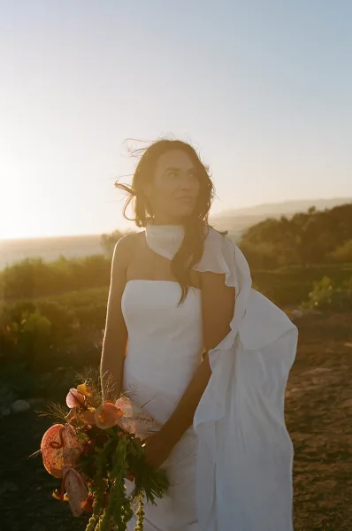 Bridal portrait of a bride in a strapless wedding dress holding anthurium bouquet, backlit at sunset on a hilltop path with greenery