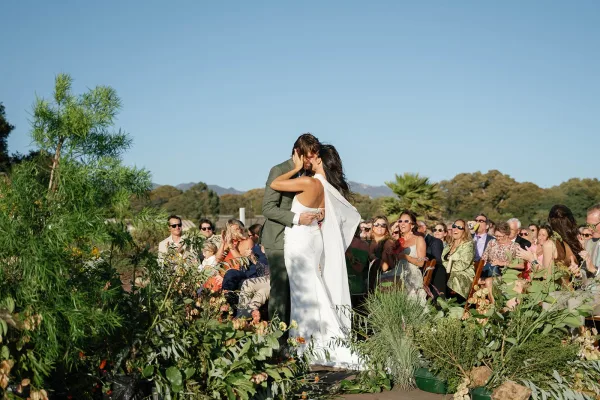 Ceremony kiss as bride in a white sheath dress and long veil kisses groom in a suit beside aisle florals in a garden setting