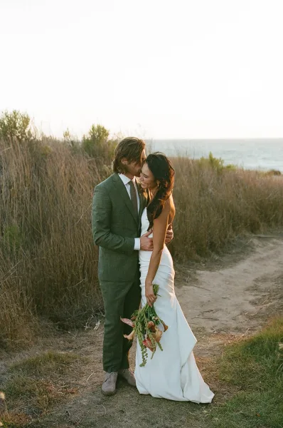Couple portrait of bride and groom embrace as he kisses her forehead, bouquet in hand on a coastal bluff above the ocean and tall grass