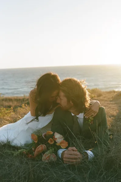 Couple portrait of bride and groom cuddling in grass, bouquet in hand, with ocean coastline and sunset sky behind them