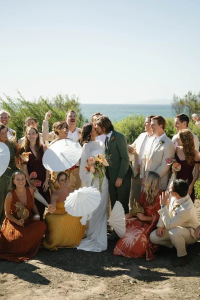 Wedding party photo with bride and groom kiss, cheering bridesmaids and groomsmen holding anthurium bouquets by the ocean view under blue sky