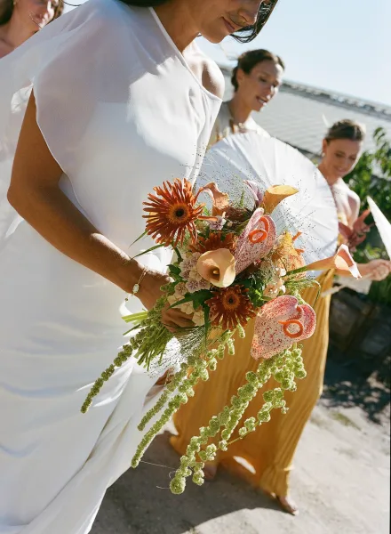 Bride walking with bouquet of calla lilies and gerbera daisies, veil flowing as bridesmaids in mustard dresses follow on an outdoor walkway