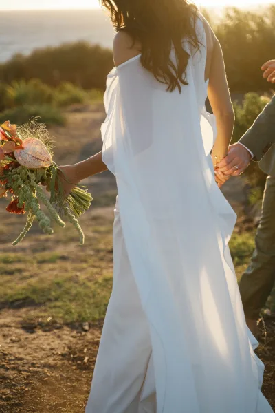 Couple portrait of bride and groom holding hands, walking away on a dirt path at sunset, her off-shoulder gown and bouquet in view