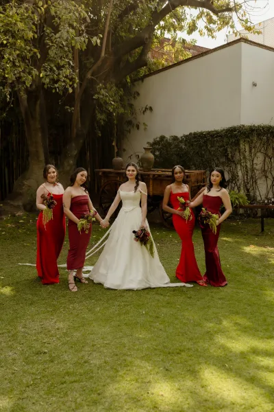 Bride and bridesmaids in a bridal party portrait, holding lush bouquets, stand by ivy-draped white wall and wooden cart in a garden.