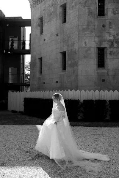 Bridal portrait of a bride with long veil in a gravel courtyard beside a stone building and picket fence, framed by neat hedges