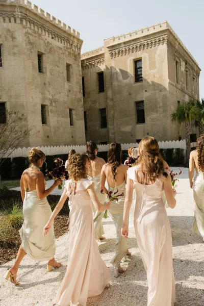 Bridesmaids walking in satin bridesmaid dresses through a courtyard beside a stone building, passing a white picket fence and a palm tree.