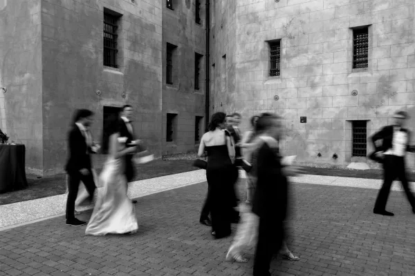 Cocktail hour guests mingle at an outdoor cocktail reception in tuxedos beside a stone courtyard with barred windows and brick pavers.