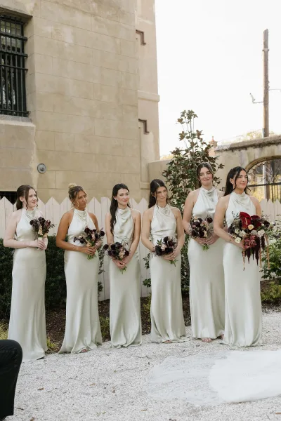 Bridesmaids portrait in satin bridesmaid dresses with cowl necklines, posed by a stone wall and white picket fence along a gravel aisle.