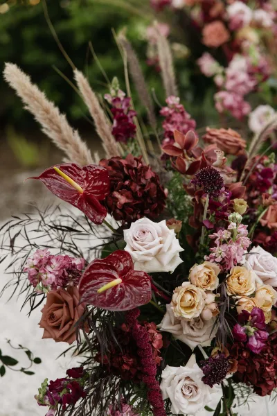 Wedding floral arrangement in burgundy and blush florals with pampas grass accents, set against lush garden greenery and a gravel path.