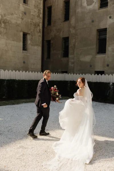 Bride and groom walking across a gravel courtyard in a candid wedding photo, her long veil trailing by a stone wall and white picket fence.