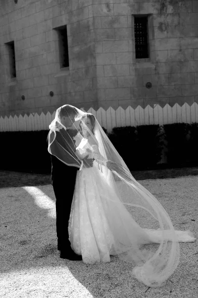 Couple portrait of a bride and groom under a sweeping veil, embraced beside a stone wall and white picket fence on a gravel path with hedges.
