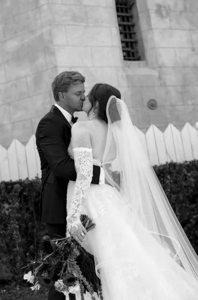 Wedding kiss portrait during an outdoor wedding portrait, groom embraces bride with a long veil beside a stone wall and hedges near a picket fence.