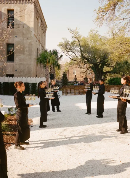 Passed champagne offered as wedding welcome drinks on serving trays in a courtyard with palm trees, a stone building, and clear sky.