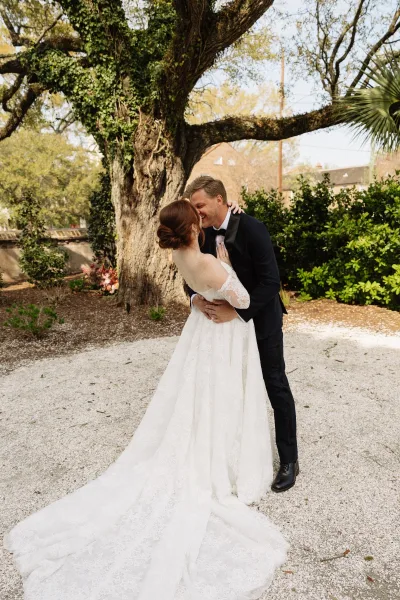 Couple portrait of a bride and groom embracing, her lace sleeves catching the light beneath a large tree amid lush garden greenery.
