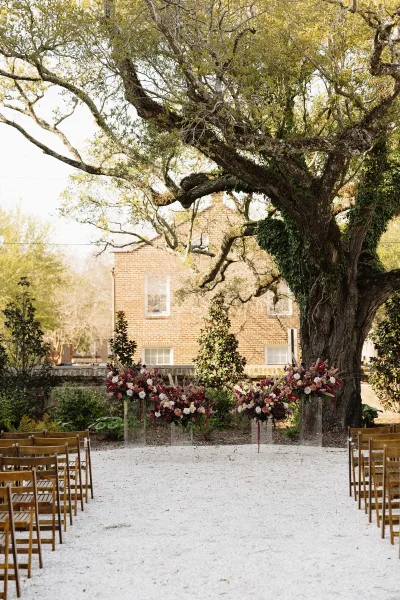 Outdoor ceremony setup for a garden wedding ceremony beneath a large tree, acrylic pedestals at the altar, a gravel aisle, and brick building.