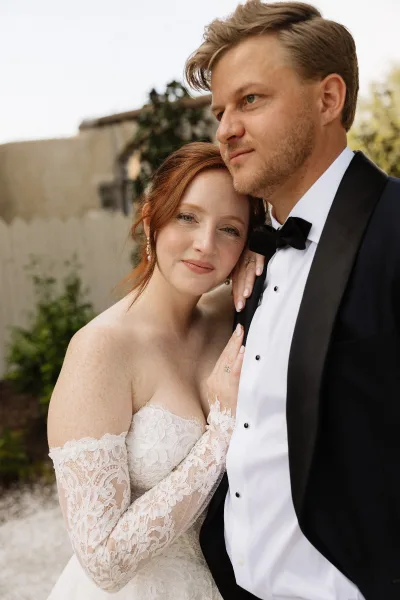 Couple portrait in a close-up of the bride and groom; she leans into him with delicate lace sleeves, framed by lush garden greenery.