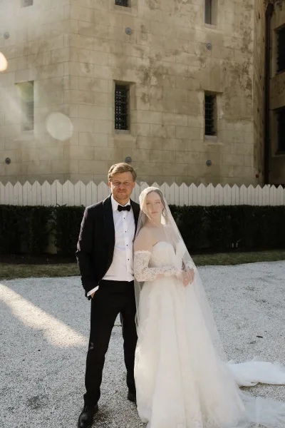Couple portrait featuring a bride with veil and her groom, framed by a stone wall and white picket fence in a quiet gravel courtyard.