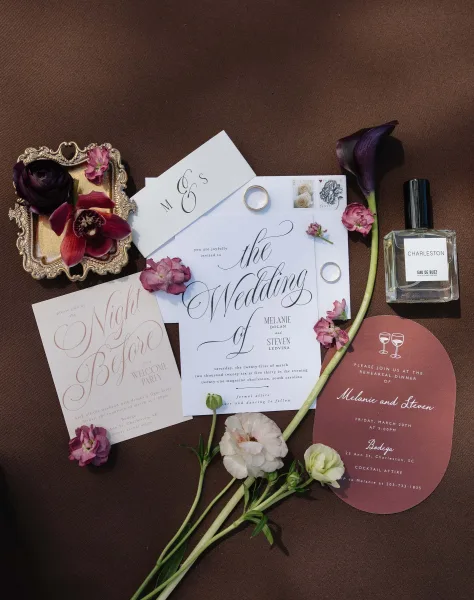 Wedding invitation suite featuring elegant calligraphy wedding invitations, accented by wedding rings on a brown fabric backdrop