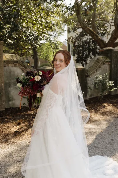 Bridal portrait of a bride with bouquet in a lace gown, standing on a gravel path by a stone wall under soft garden trees.