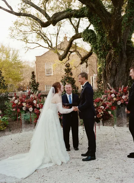 Ceremony moment of the couple's vow exchange, hands joined beneath a large garden tree, bride’s long veil flowing, brick building behind.