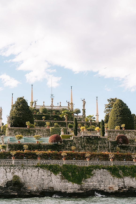 Garden venue exterior with stone balustrades and statues on a formal terrace overlooking a lake under a cloudy sky, framed by topiary