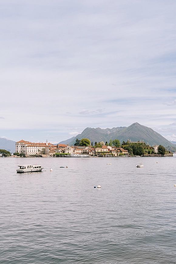 Lake wedding venue and lakeside wedding location overlooking a calm lake with boats and buoys, shoreline village, mountains, and cloudy sky