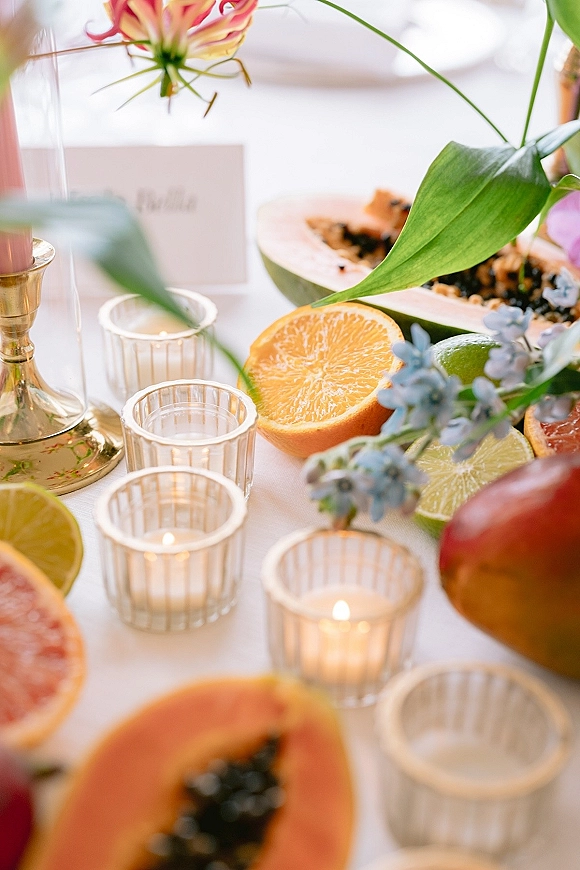 Reception tablescape with citrus wedding tablescape accents of halved oranges, limes, grapefruit and papaya, plus flowers and votive candles on white linen