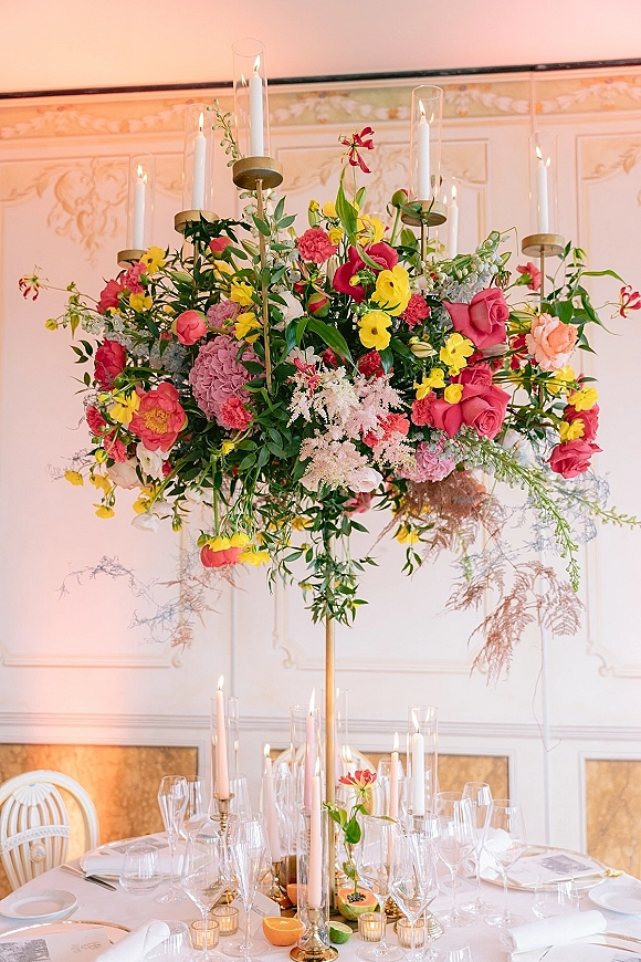 Reception centerpiece with tall floral centerpiece on a gold stand, surrounded by taper candles and glass hurricanes on a set table indoors