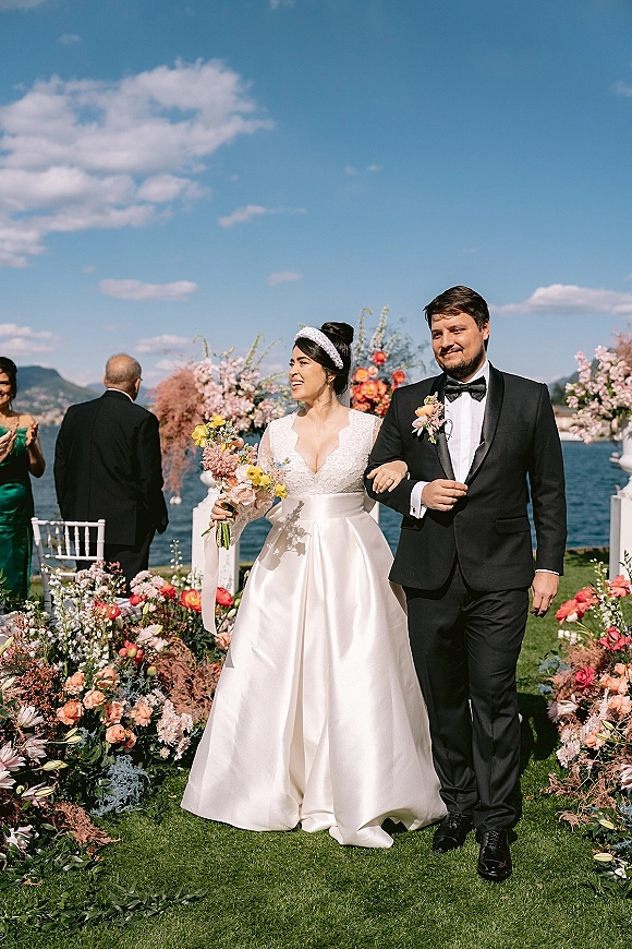 Wedding recessional as bride and groom walk arm in arm down the aisle, she holds a bouquet; lake and mountains under blue sky behind
