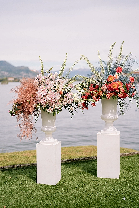 Wedding floral arrangement with ceremony entrance florals in white urn vases on pedestals, pink and red blooms beside a lakeside lawn with mountains