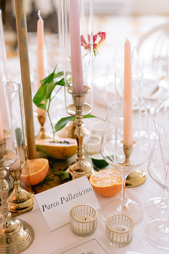 Reception tablescape with wedding table decor featuring pastel taper candles in glass hurricane holders, citrus, greenery, and place cards on white linen